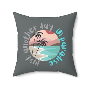 May include: Gray throw pillow with a circular design featuring palm trees, a beach, and ocean waves. The text "Just another day in paradise" is written around the circle.