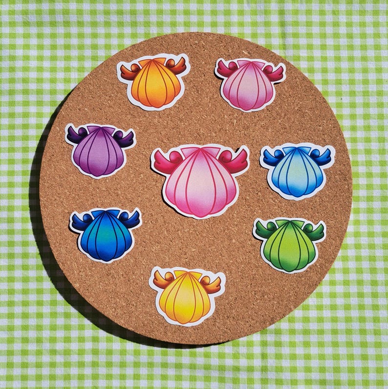 Op de afbeelding: Een verzameling kleurrijke schelpenstickers gerangschikt op een ronde kurken plank. De stickers tonen verschillende tinten roze, blauw, geel, paars en groen. Elke schelp heeft een witte omtrek en decoratieve vleugelachtige accenten.