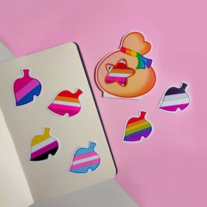 Könnte beinhalten: Ein Set mit zehn bunten, blattartigen Aufklebern mit Regenbogenstreifen. Die Aufkleber zeigen verschiedene Farbkombinationen aus Rot, Orange, Gelb, Grün, Blau, Indigo und Violett. Die Aufkleber sind auf einem rosa und weißen Hintergrund angeordnet.