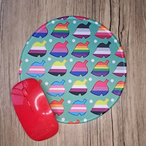 Op de afbeelding: Een ronde muismat met een teal achtergrond en witte stippen. De mat is versierd met een herhalend patroon van gestileerde bladeren in verschillende kleuren die verschillende pride-vlaggen vertegenwoordigen.