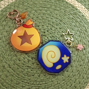 May include: Two Animal Crossing themed keychains. One is a gold coin bag with a brown star and the other is a blue shell with a yellow spiral and a white star.