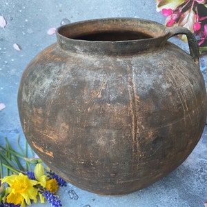 May include: A large, brown, earthenware pot with a single handle. The pot has a rough, textured surface and appears to be very old.