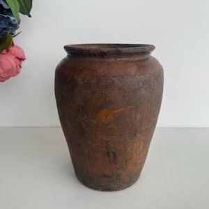 Può includere: Un vaso in ceramica marrone con una base arrotondata e una parte superiore leggermente svasata. Il vaso ha un aspetto rustico e invecchiato con graffi e imperfezioni visibili.