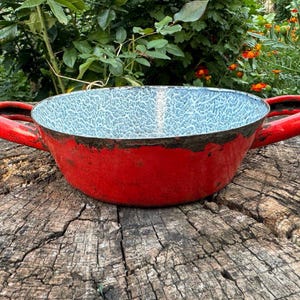 May include: A red enamelware bowl with two handles. The bowl has a white speckled interior and a dark rim.