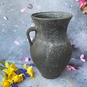 Könnte beinhalten: Eine schwarze Keramikvase mit einer breiten Öffnung und einem Henkel. Die Vase hat ein rustikales, gealtertes Aussehen und ist perfekt für die Präsentation von Blumen.