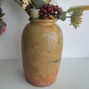 May include: A vintage, rustic, glazed ceramic vase with a light brown, yellow, and orange colour scheme. The vase has a round, wide base and a narrow opening. It is filled with a bouquet of dried flowers and berries.