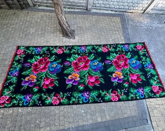 May include: A rectangular, black textile featuring a colourful floral design. Large pink and blue flowers are set amongst smaller blooms and green leaves. The edges are trimmed with red fringe.
