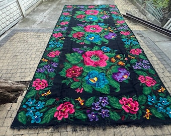 May include: A large, rectangular black rug with a vibrant floral pattern. The design features large pink, blue, and purple flowers with green leaves. The rug has a fringed edge and is displayed outdoors.