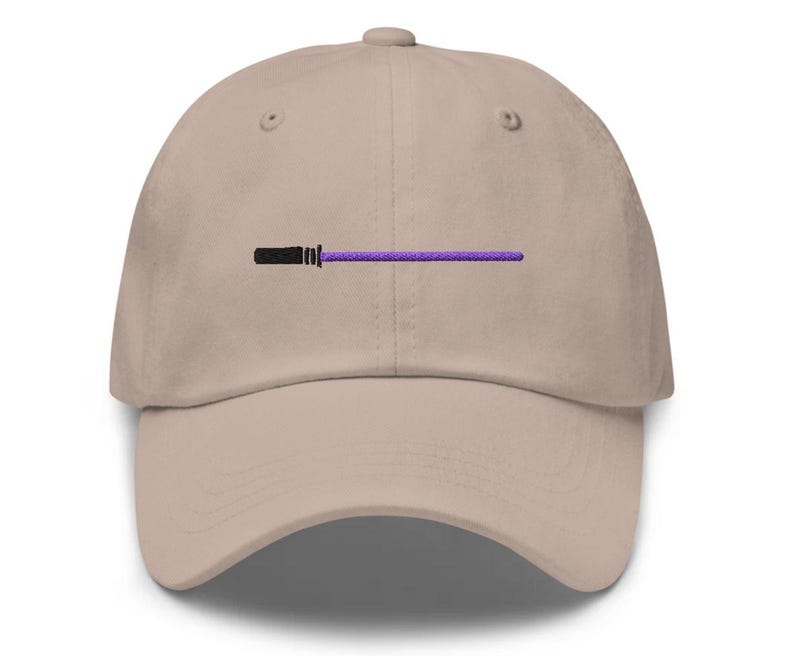 Puede incluir: Gorra de b&eacute;isbol beige con un dise&ntilde;o de sable de luz negro y morado bordado en la parte delantera. La gorra tiene una visera curva y un cierre de correa de tela en la parte trasera.