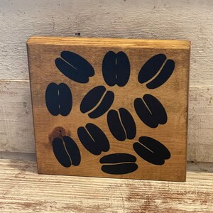 May include: A wooden sign with a brown background and black coffee bean silhouettes. The sign has a rustic look and is perfect for a coffee lover's home.