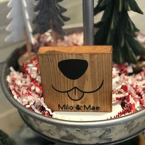 May include: Wooden block with a dog face design and the text "Milo & Mae" on a tiered tray with red and white shredded paper and small artificial Christmas trees.