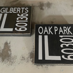 May include: Two black wooden signs with white lettering. The top sign says "GILBERTS IL 60136" and the bottom sign says "OAK PARK IL 60301".