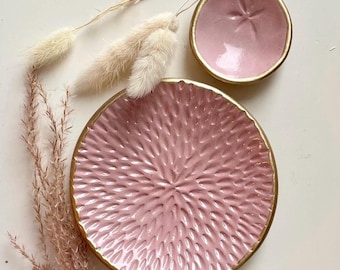 May include: A set of two round ceramic dishes in a soft pink glaze with gold-painted rims. The larger dish features a textured, radiating pattern, while the smaller dish has a smooth surface with a subtle, embossed star-like center detail.