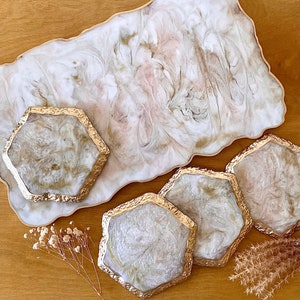 May include: Set of four white and gold hexagon-shaped coasters with a marbled design. The coasters are on a larger white and gold marbled tray.