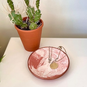 May include: A pink and white marbled ceramic dish with a gold chain necklace and a round pendant resting inside. The dish is on a white surface next to a potted cactus.