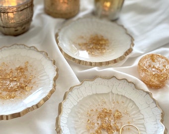 May include: Three white resin ring dishes with gold leaf accents and a gold rim. The dishes are on a white surface.