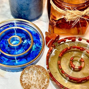 May include: Three resin coasters with different designs. One coaster is blue with gold accents and a concentric circle pattern. Another coaster is clear with a red concentric circle pattern. The third coaster is clear with gold glitter.