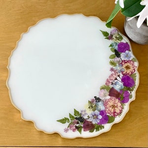 May include: A white round resin tray with a scalloped edge and a border of pressed flowers in shades of purple, pink, and white.