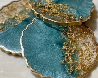 May include: Three teal blue resin coasters with gold flecks and a gold metal rim. The coasters are in a stack with the top coaster slightly offset.
