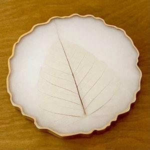 May include: A white resin coaster with a gold rim and a delicate pressed leaf design.