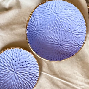 May include: Two decorative bowls with a light purple glaze and a gold rim. The bowls have a textured, raised design that resembles a flower.