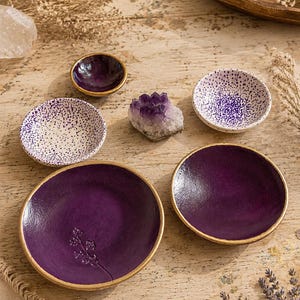 May include: A collection of five small ceramic dishes in varying sizes, featuring deep purple and white speckled finishes with gold-painted rims. One dish displays a delicate embossed floral sprig, while a raw amethyst crystal sits centrally.