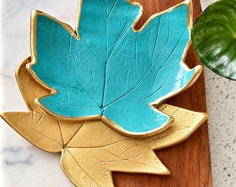 May include: Two leaf-shaped decorative dishes stacked together. The top dish is a vibrant turquoise color with gold-painted edges, while the bottom dish is entirely gold. Both pieces feature detailed, realistic vein textures across their surfaces.