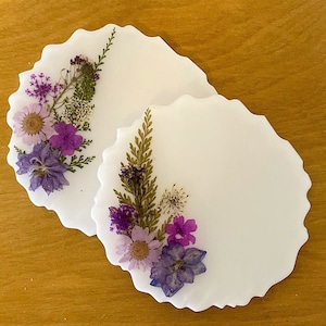 May include: Two white coasters with a scalloped edge, each decorated with pressed flowers and greenery. The flowers are purple, white, and blue.