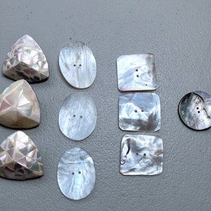 May include: Assortment of white mother of pearl buttons in various shapes and sizes, including oval, square, and triangular. Some buttons have a decorative pattern.