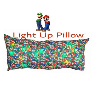 May include: A long rectangular pillow with a light-up feature. The pillow is covered in a blue, green, and red fabric with a repeating pattern of Mario and Luigi characters. The text "Light Up Pillow" is written in red letters above the pillow.