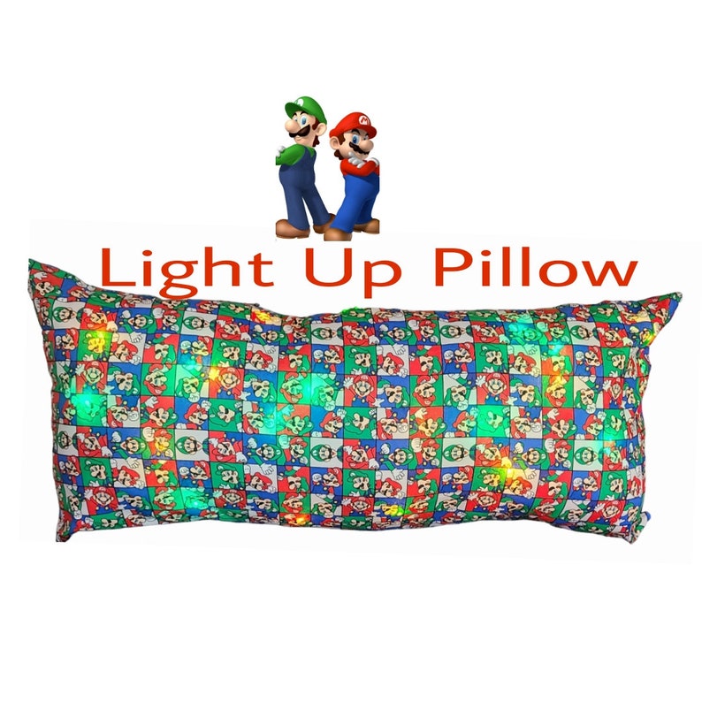 May include: A long rectangular pillow with a light-up feature. The pillow is covered in a blue, green, and red fabric with a repeating pattern of Mario and Luigi characters. The text "Light Up Pillow" is written in red letters above the pillow.