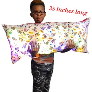 May include: A white and colorful pillow with a cartoon design and built-in LED lights. The pillow is 35 inches long and is being held by a person.