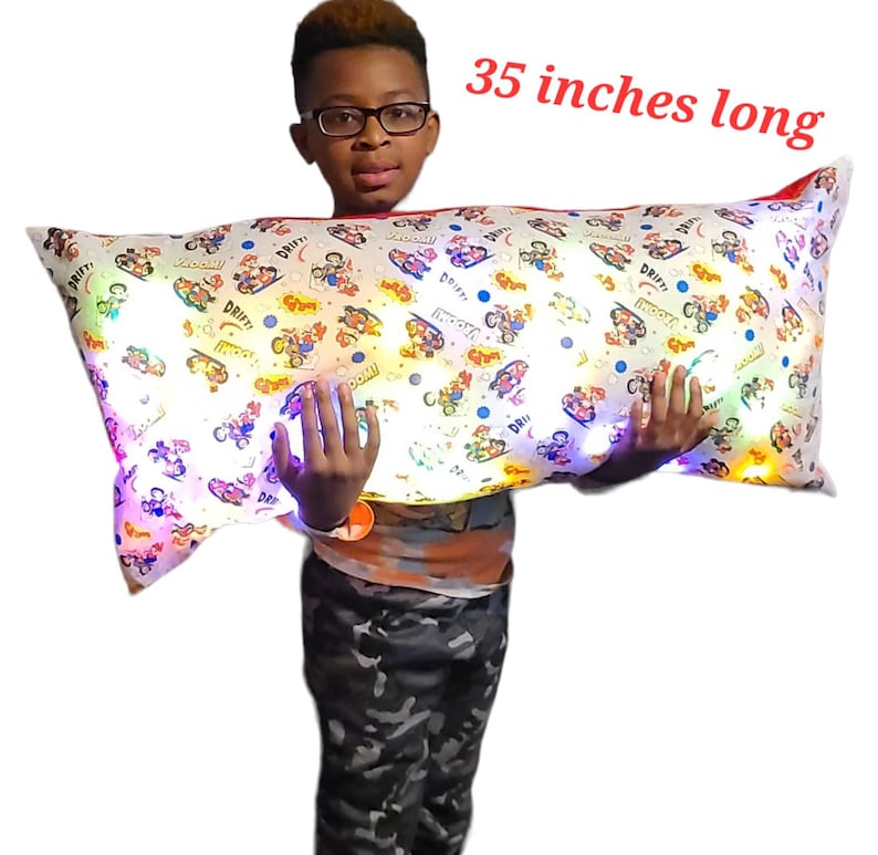 May include: A white and colorful pillow with a cartoon design and built-in LED lights. The pillow is 35 inches long and is being held by a person.
