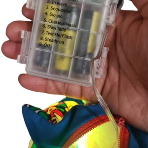 May include: A clear plastic battery pack with a list of light modes: Combination, In wave, Sequential, Slo glo, Chasing/Flash, Slow fade, Twinkle/Flash, Steady on, Off. The battery pack is connected to a blue and red fabric with a yellow light shining through.