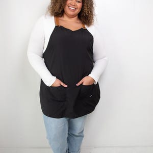 Pu&ograve; includere: Una donna indossa un grembiule nero con cinturini in pelle marrone e una camicia bianca a maniche lunghe. Indossa jeans blu e scarpe da ginnastica bianche.