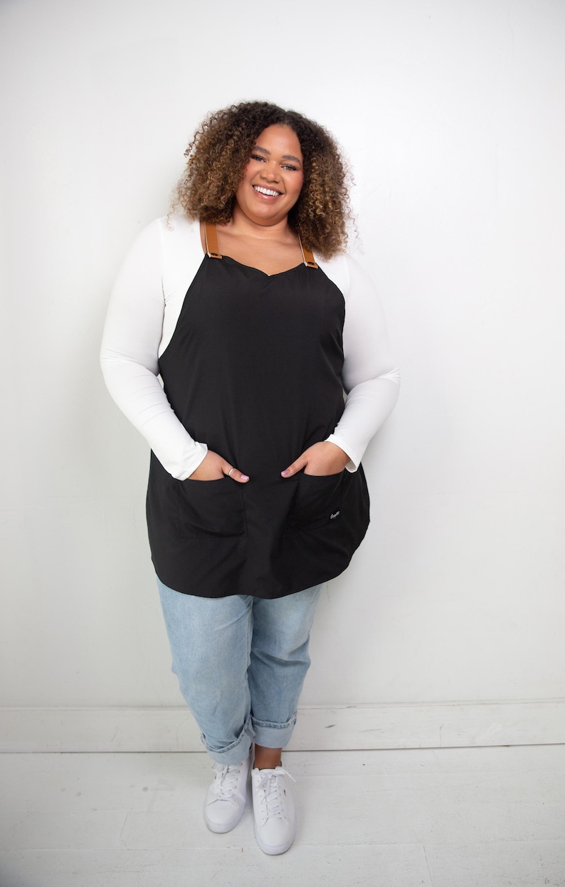 Pu&ograve; includere: Una donna indossa un grembiule nero con cinturini in pelle marrone e una camicia bianca a maniche lunghe. Indossa jeans blu e scarpe da ginnastica bianche.