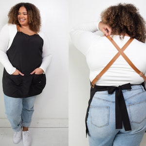 Pu&ograve; includere: Un grembiule nero con due tasche e cinturini regolabili in pelle marrone. Il grembiule &egrave; indossato sopra una camicia bianca a maniche lunghe e jeans blu.