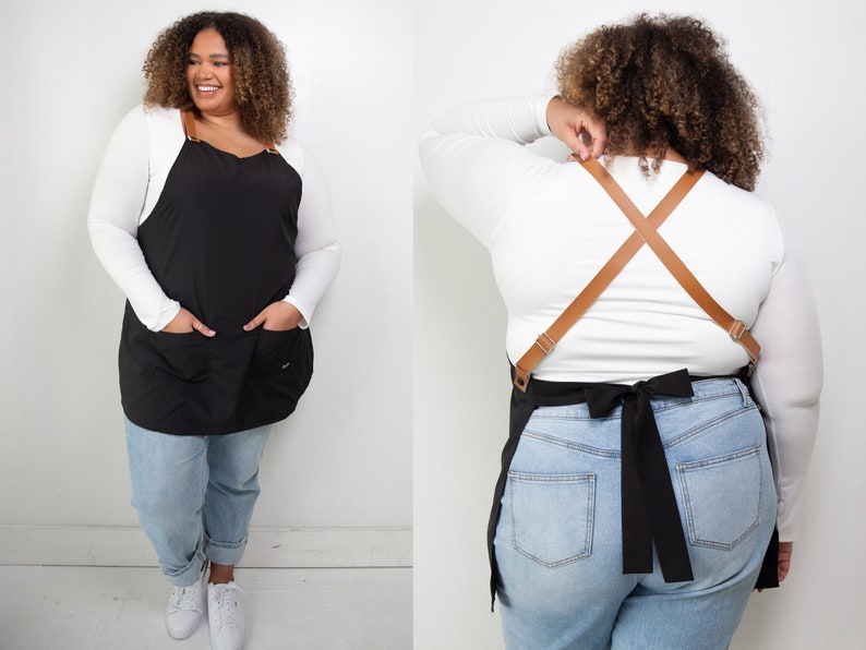 Pu&ograve; includere: Un grembiule nero con due tasche e cinturini regolabili in pelle marrone. Il grembiule &egrave; indossato sopra una camicia bianca a maniche lunghe e jeans blu.