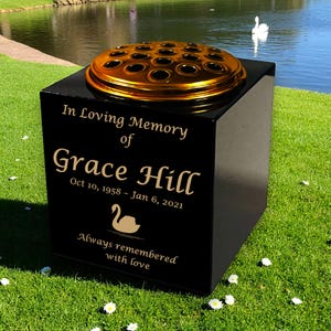 May include: A black memorial vase with a gold-coloured flower grid on top. The vase has the inscription "In Loving Memory of Grace Hill Oct 10, 1958 - Jan 6, 2021" and a swan graphic. The text "Always remembered with love" is also included.