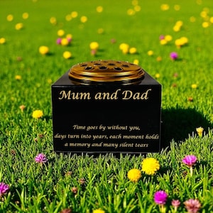 May include: A black memorial vase inscribed with "Mum and Dad" in gold. It features a gold-coloured top for flowers. The text "Time goes by without you..." is below. The vase is placed in a grassy area with colourful flowers.