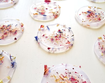 Peut inclure: Un ensemble de sous-verres ronds en résine transparente avec des pétales de fleurs séchées et des paillettes dorées incrustés. Les sous-verres sont disposés en rangée sur une surface blanche.