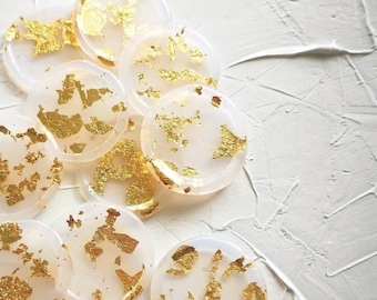 Peut inclure: Un ensemble de dessous-de-verre ronds, blancs avec des accents de feuilles d'or. Les dessous-de-verre sont disposés en pile sur une surface texturée blanche.