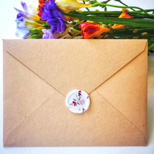 Peut inclure: Une enveloppe en papier brun avec un sceau de cire blanc orn&eacute; d'un motif floral. L'enveloppe est pos&eacute;e sur une surface blanche avec un bouquet de fleurs color&eacute;es en arri&egrave;re-plan.