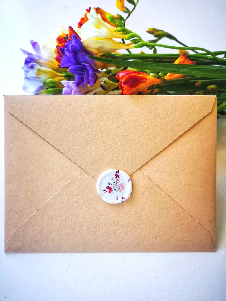 Peut inclure: Une enveloppe en papier brun avec un sceau de cire blanc orn&eacute; d'un motif floral. L'enveloppe est pos&eacute;e sur une surface blanche avec un bouquet de fleurs color&eacute;es en arri&egrave;re-plan.