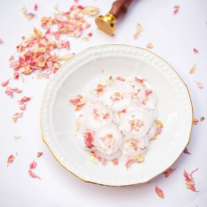 Peut inclure: Une assiette en c&eacute;ramique blanche avec un bord dor&eacute; contient un tas de sceaux de cire ronds et blancs avec des p&eacute;tales de fleurs roses &eacute;parpill&eacute;s autour. Un tampon de sceau de cire en bois et or se trouve sur une surface en marbre blanc.