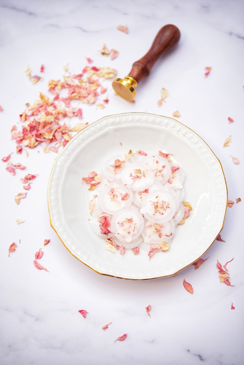 Peut inclure: Une assiette en c&eacute;ramique blanche avec un bord dor&eacute; contient un tas de sceaux de cire ronds et blancs avec des p&eacute;tales de fleurs roses &eacute;parpill&eacute;s autour. Un tampon de sceau de cire en bois et or se trouve sur une surface en marbre blanc.