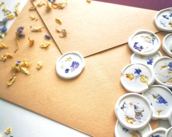 Peut inclure: Gros plan sur une enveloppe beige avec un sceau de cire blanc. Le sceau présente un motif de fleurs séchées dans des tons bleus et dorés. L'enveloppe est entourée de pétales de fleurs séchées.