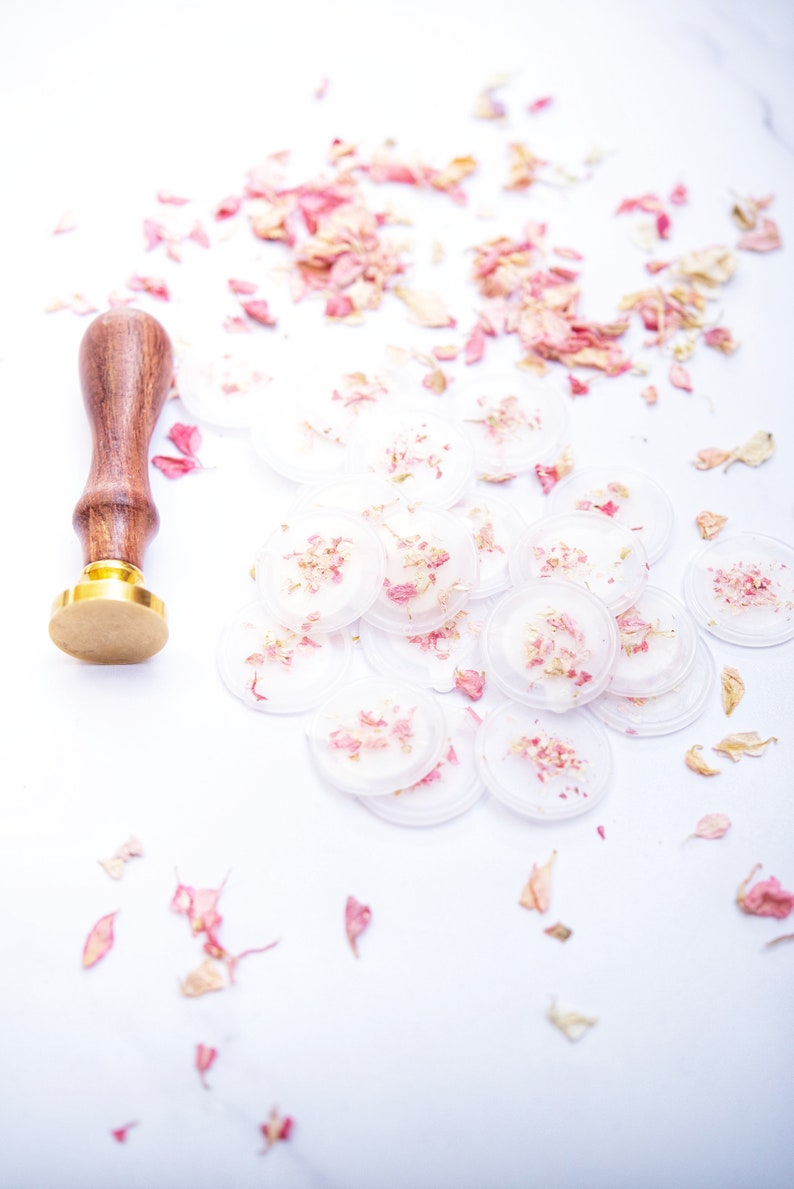 Peut inclure: Un ensemble de sceaux ronds en cire transparente avec des p&eacute;tales de fleurs roses s&eacute;ch&eacute;es incrust&eacute;s &agrave; l'int&eacute;rieur. Les sceaux sont &eacute;parpill&eacute;s sur une surface blanche &agrave; c&ocirc;t&eacute; d'un tampon &agrave; cire en bois et en or.