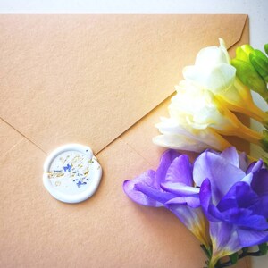 Peut inclure: Une enveloppe brune avec un sceau de cire blanc et un bouquet de freesia blanches et violettes.