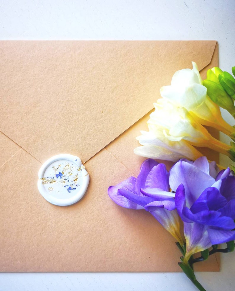 Peut inclure: Une enveloppe brune avec un sceau de cire blanc et un bouquet de freesia blanches et violettes.